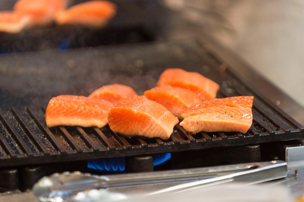 Pavés de saumon posés sur la grille en fonte d'un barbecue, flammes visibles sous la grille, une pince à portée de main